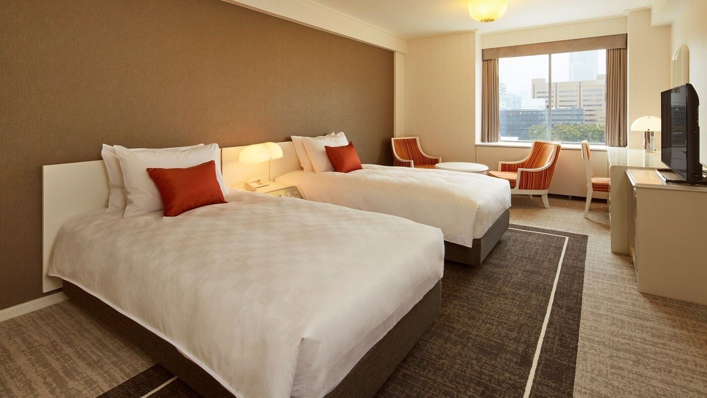 A hotel room with two beds, a desk, a chair, and a TV.