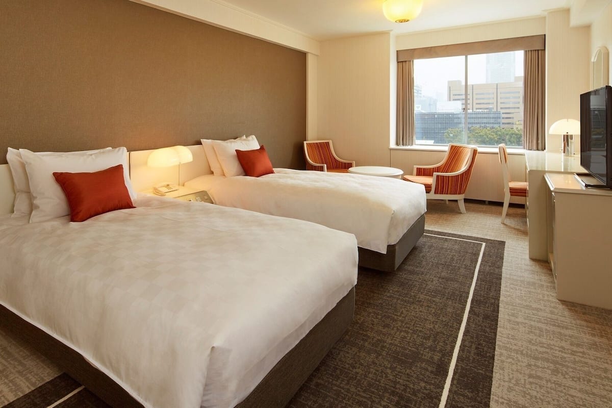 A hotel room with two beds, a desk, a chair, and a TV.