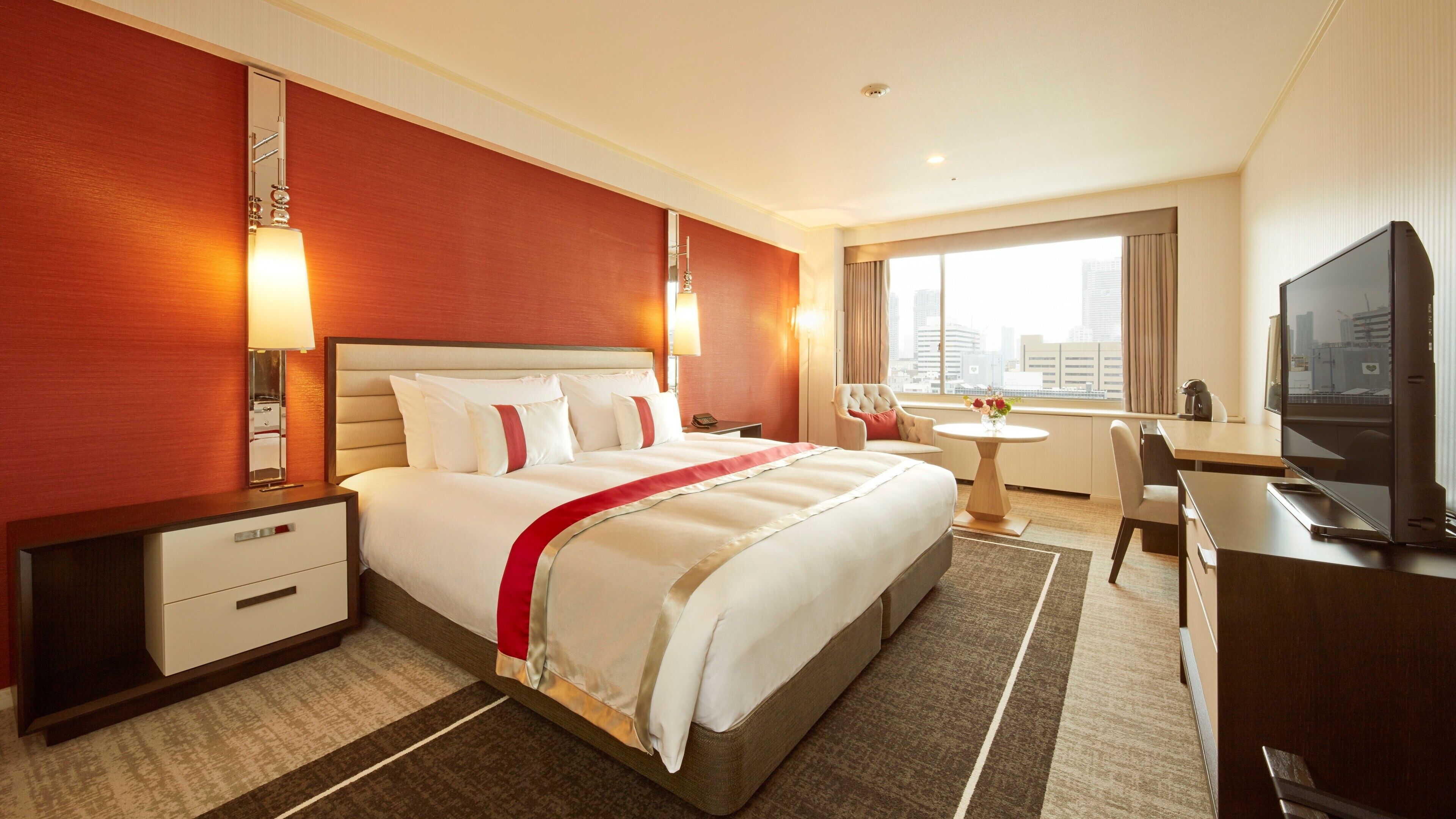 A hotel room with a large bed, bedside tables, a desk, a chair, a television, and a window with a city view.