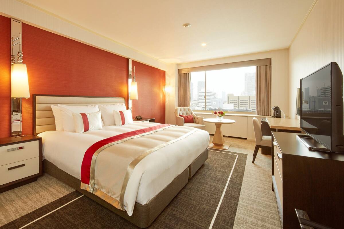 A hotel room with a large bed, bedside tables, a desk, a chair, a television, and a window with a city view.