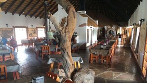 A restaurant with wooden tables and chairs, a large tree trunk centerpiece, stone flooring, and a high ceiling with wooden beams. - Canon Village (Fish River Canyon)