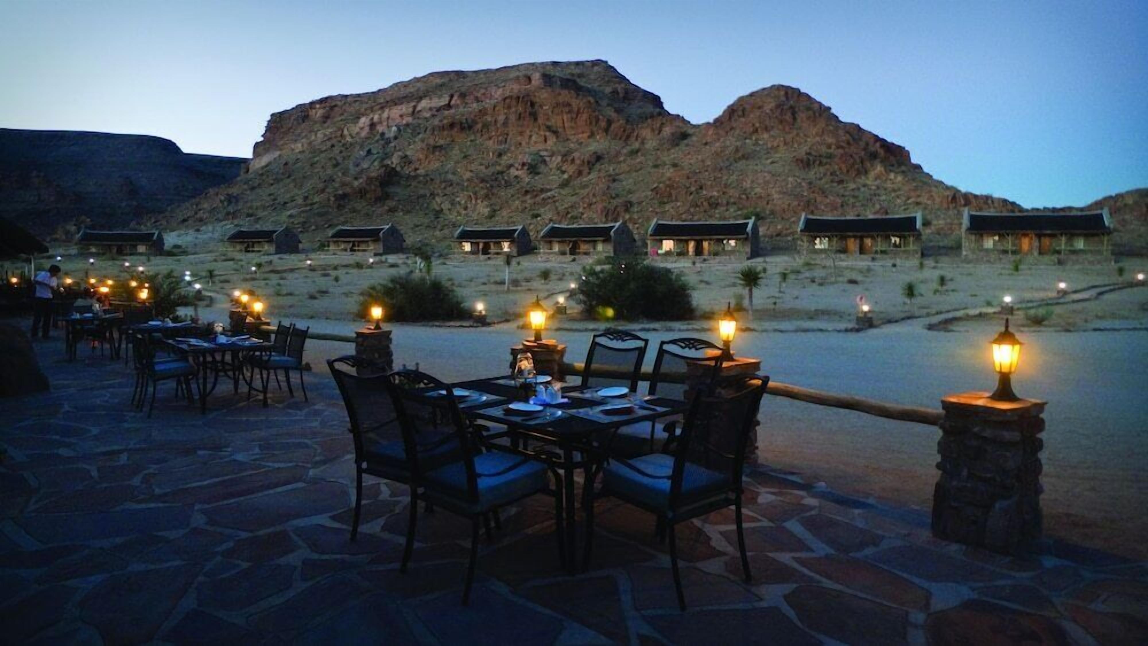 Un área de comedor al aire libre con mesas y sillas, piso de piedra y vista a un paisaje montañoso.