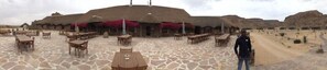 Enjoy al fresco dining in a serene desert setting, surrounded by a thatched-roof building and sparse vegetation.