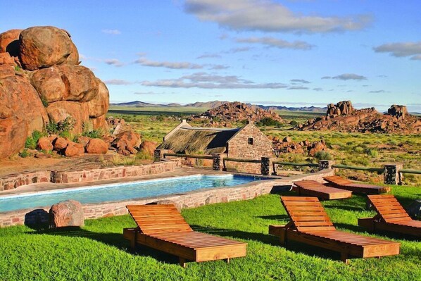 Relax by the serene pool surrounded by lush greenery and boulders, with a rustic building in the background.