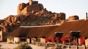 A stone building with a thatched roof, surrounded by a rocky landscape and red flowering plants. - Canon Village (Fish River Canyon)