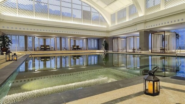 A large indoor swimming pool with a glass barrier, surrounded by seating areas and a high ceiling with a lattice design. - Beijing Grand Hotel (Beijing)