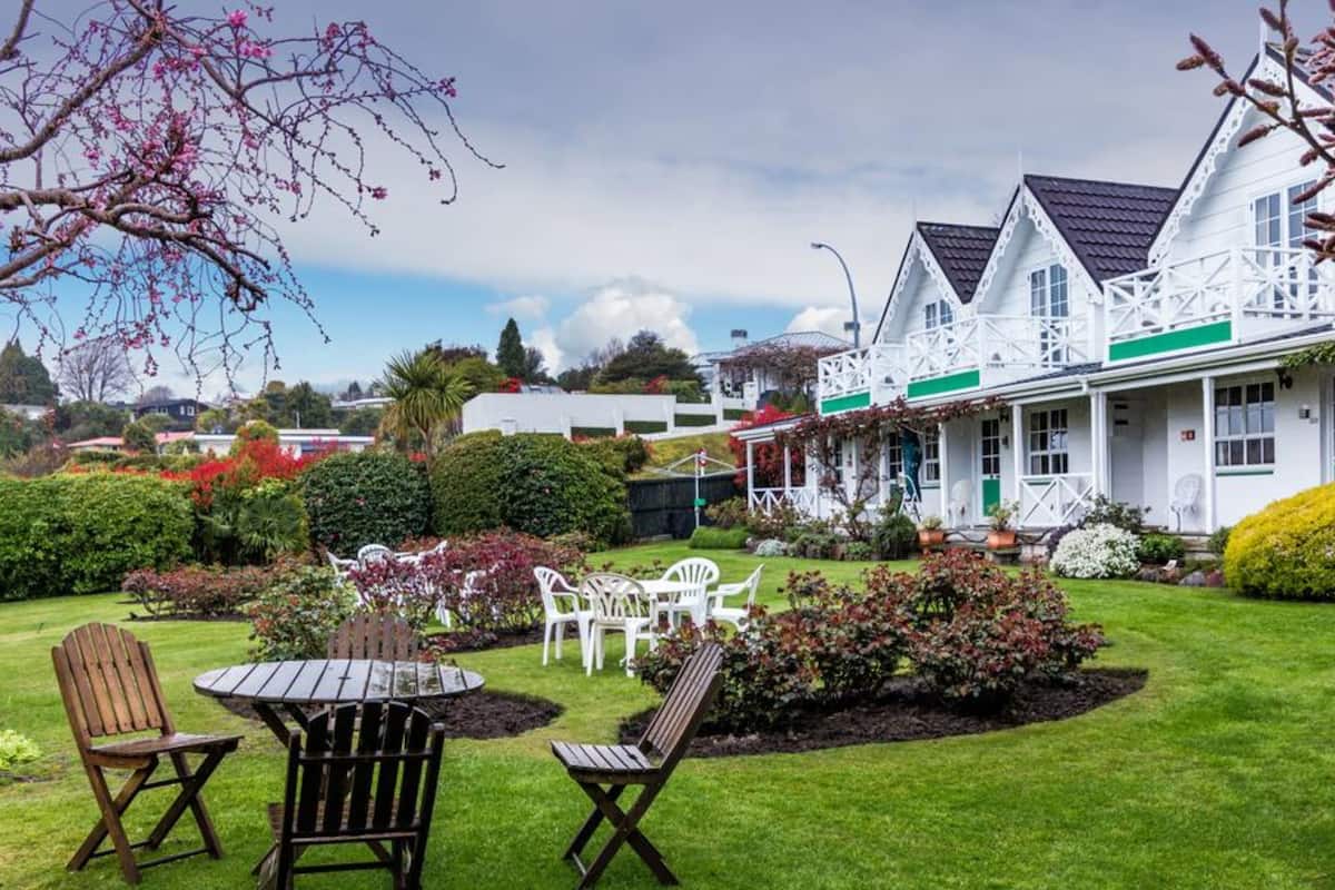 A white building with a green roof, surrounded by a well-maintained garden with various plants and a lawn. There are outdoor seating arrangements with chairs and a table.
