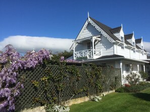 Una casa blanca de dos pisos con techo inclinado, un balcón y una cerca de madera adornada con flores moradas.
