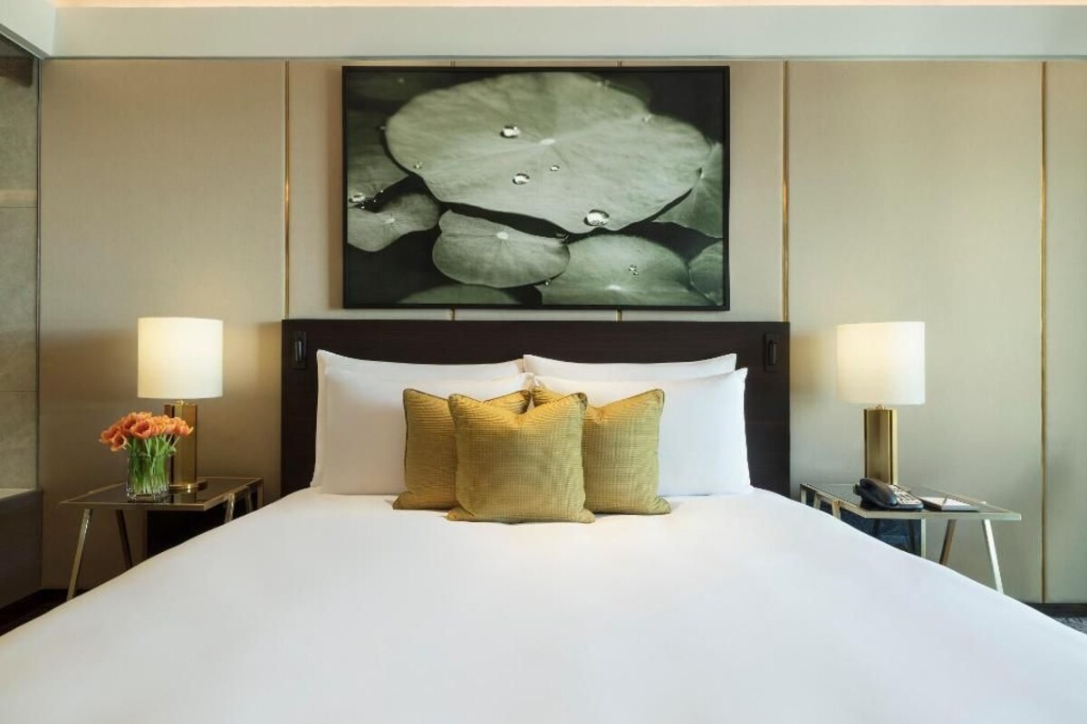 Habitación de hotel con una cama grande, dos lámparas de noche, un florero con flores y una obra de arte en la pared con nenúfares.