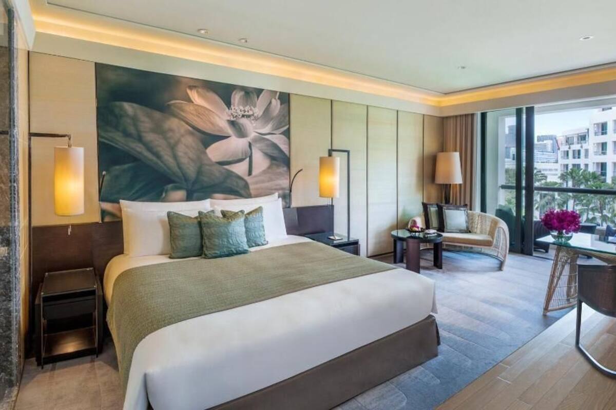 A modern hotel room with a large bed, a sitting area with a chair and table, a view of buildings outside, and a large floral artwork on the wall.