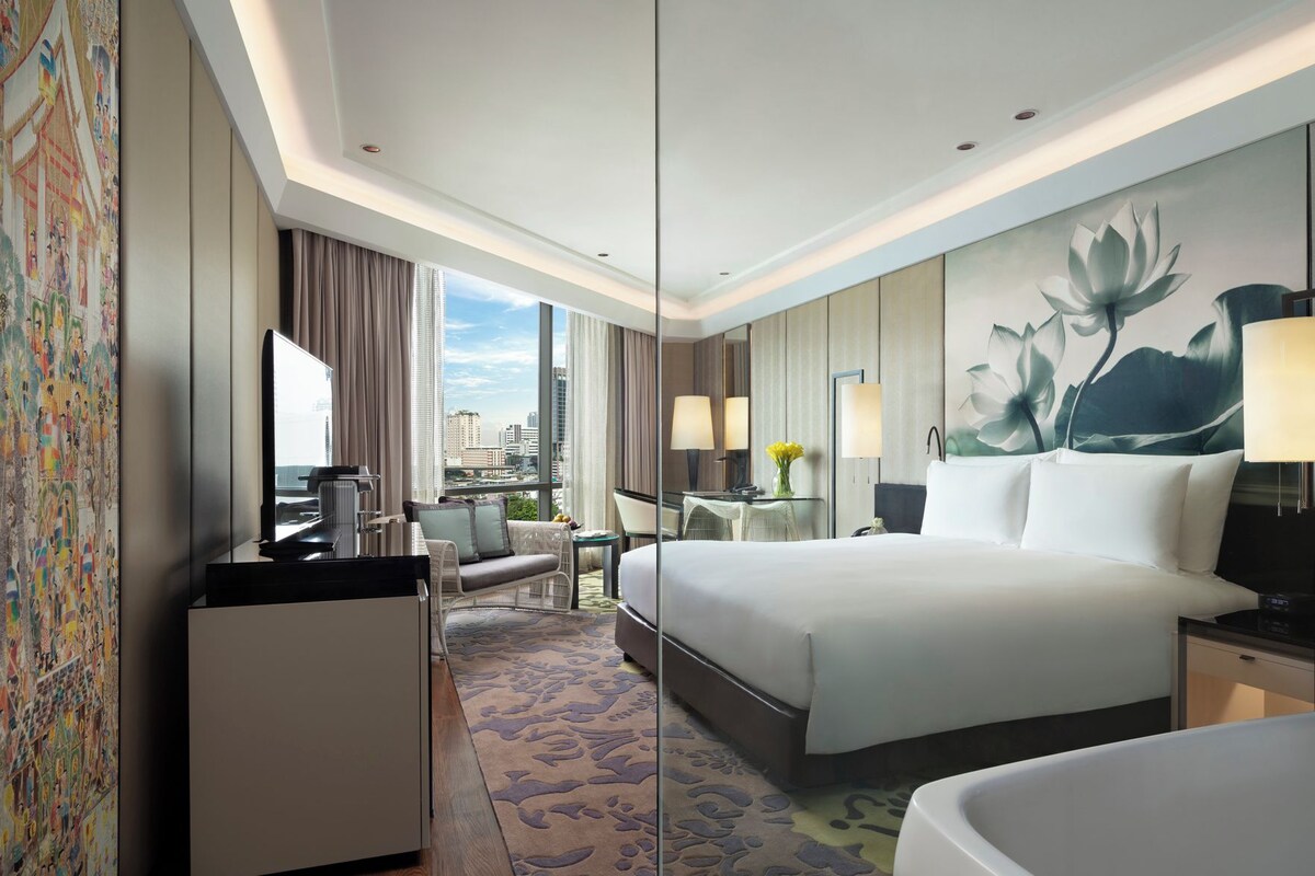 Habitación de hotel con cama, escritorio, silla y un ventanal con vista a la ciudad.
