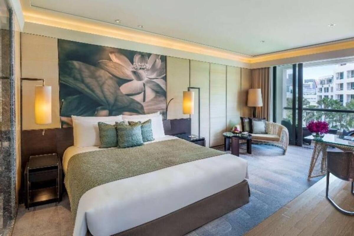 A hotel room with a large bed, a sitting area with a chair and table, a large window with a view, and a decorative wall painting.
