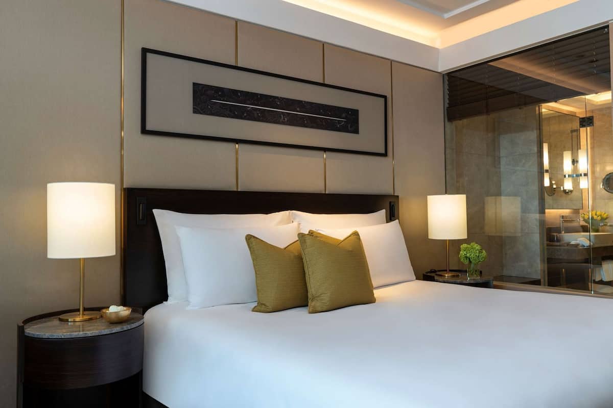 A hotel room with a large bed, two bedside tables, a nightstand, a framed artwork above the bed, and a bathroom visible through a glass partition.