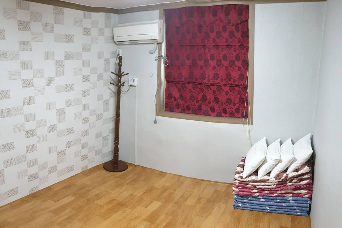 Una habitación con piso de madera, aire acondicionado montado en la pared, perchero, ventana con cortinas rojas y una pila de almohadas.