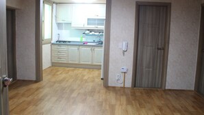 Una habitación con piso de madera, una puerta y vista a una cocina con alacenas blancas y un ventanal.