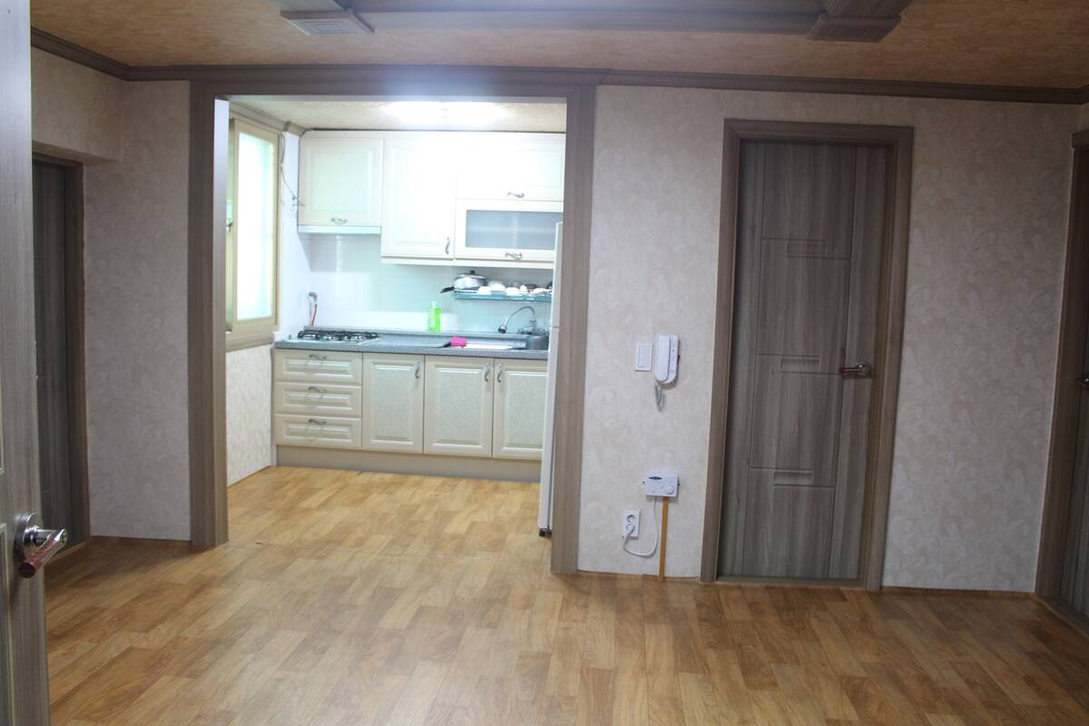 Una habitación con piso de madera, una puerta y vista a una cocina con alacenas blancas y un ventanal.