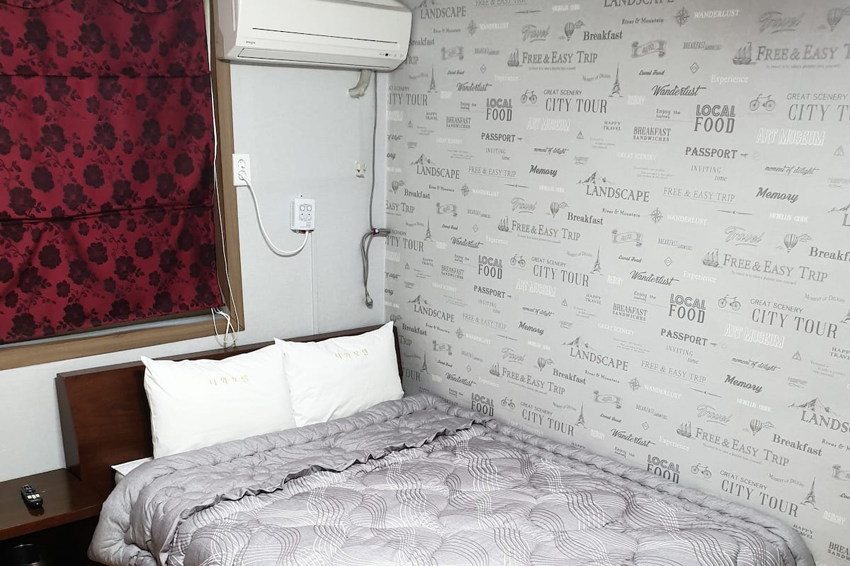 Una cama bien hecha con una colcha estampada, una mesita de noche de madera con un control remoto y una pared decorada con un patrón repetitivo de palabras relacionadas con viajes.