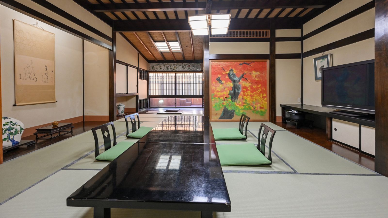 A tatami mat room with a long table, chairs, a painting, and a television.