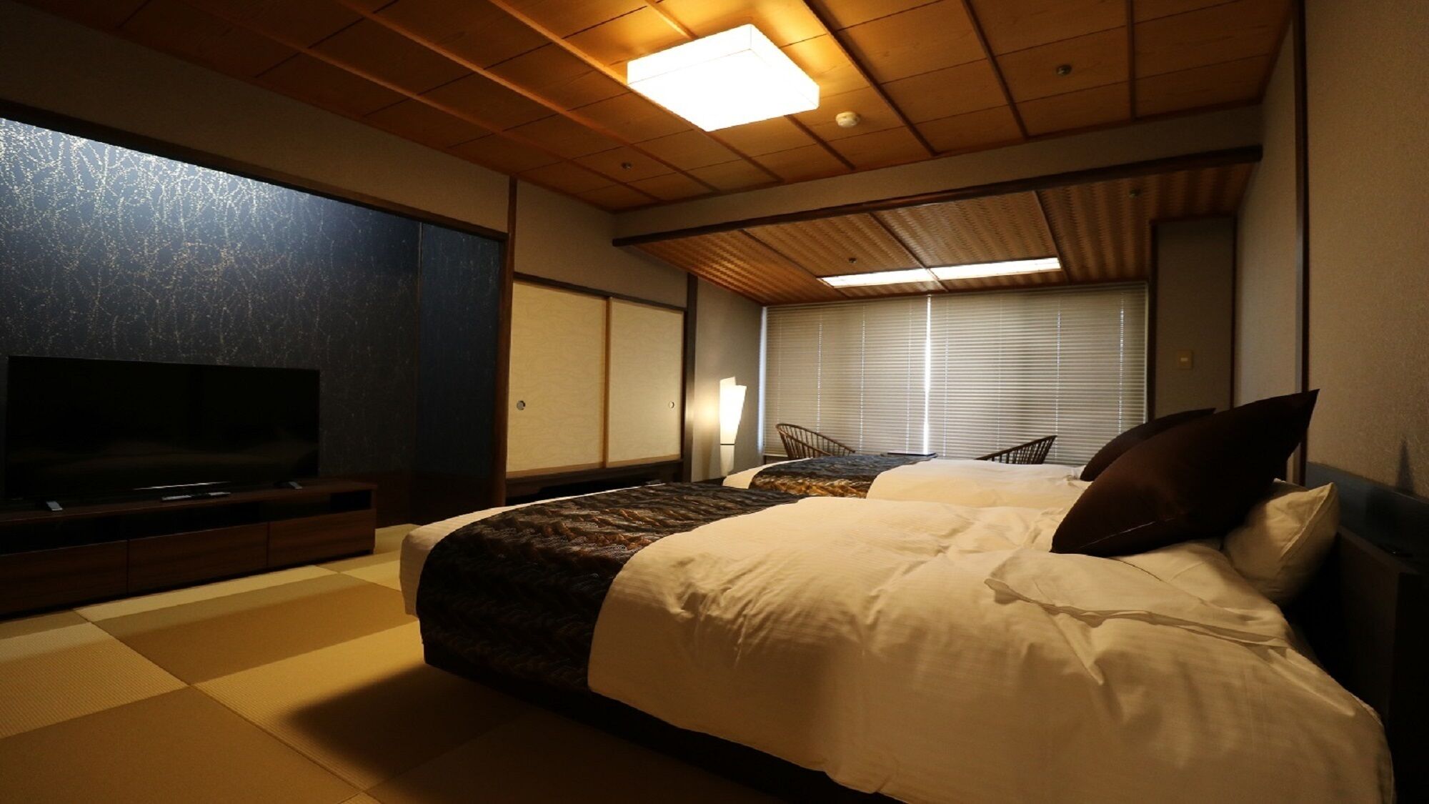 A hotel room with a large bed, a television, a window with blinds, and a wooden ceiling.