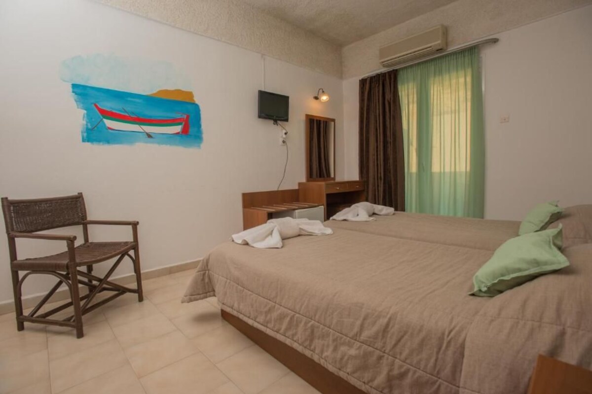 Quarto de hotel com uma cama, uma cadeira, uma escrivaninha com telefone, um quadro de um barco e uma janela com cortinas.