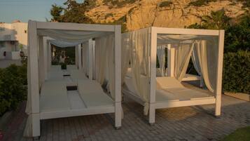 Two white, open-air beds with draped fabric, set on a paved area with a mountainous backdrop.