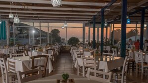 En rummelig restaurant med flere borde og stole, store vinduer der giver udsigt til solnedgangen, og hængende pendellamper.