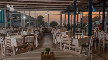Een ruime restaurantruimte met meerdere tafels en stoelen, grote ramen met uitzicht op de zonsondergang en hangende吊灯.