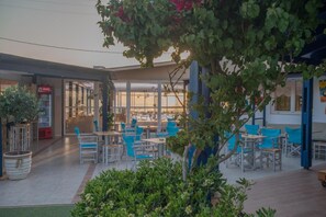 An outdoor seating area with tables and chairs, a Coca-Cola refrigerator, and a view of the beach through large windows. - Fragiskos (Faistos)