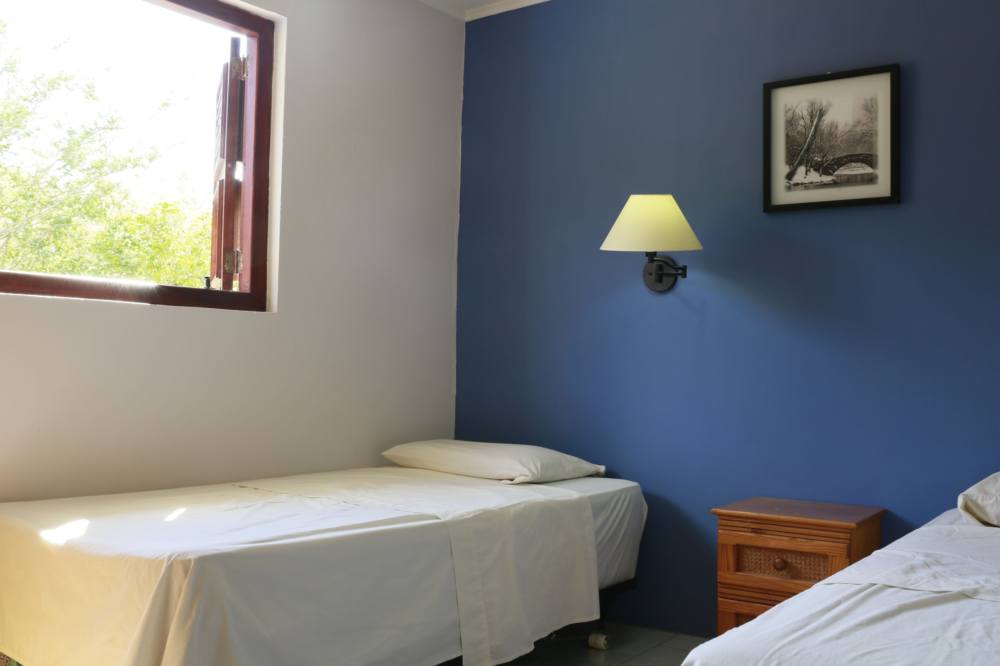 Une chambre avec un lit simple, une table de chevet, une lampe fixée au mur et un tableau encadré.