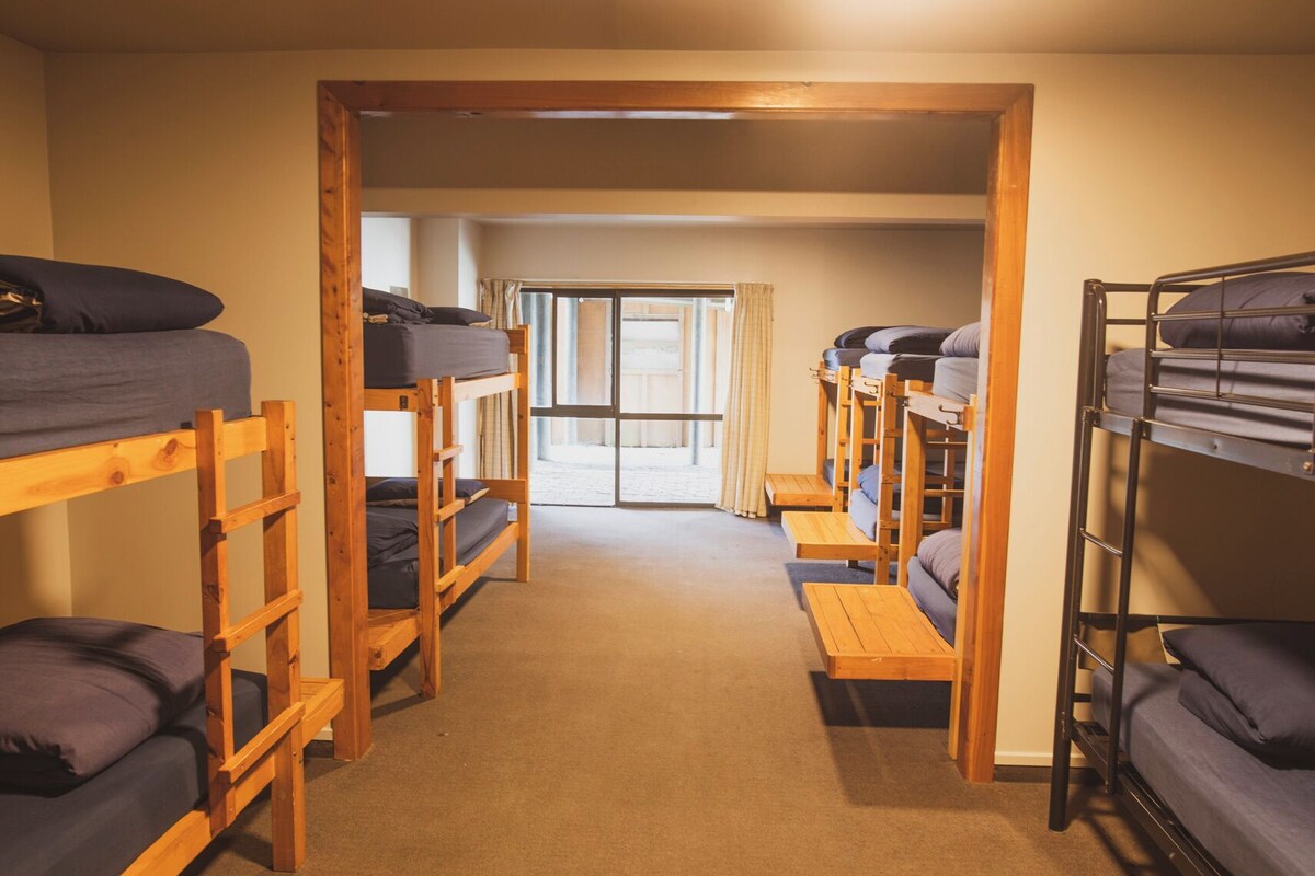 A dormitory room with bunk beds arranged in two rows, a carpeted floor, and a door leading to a bright room with a view.