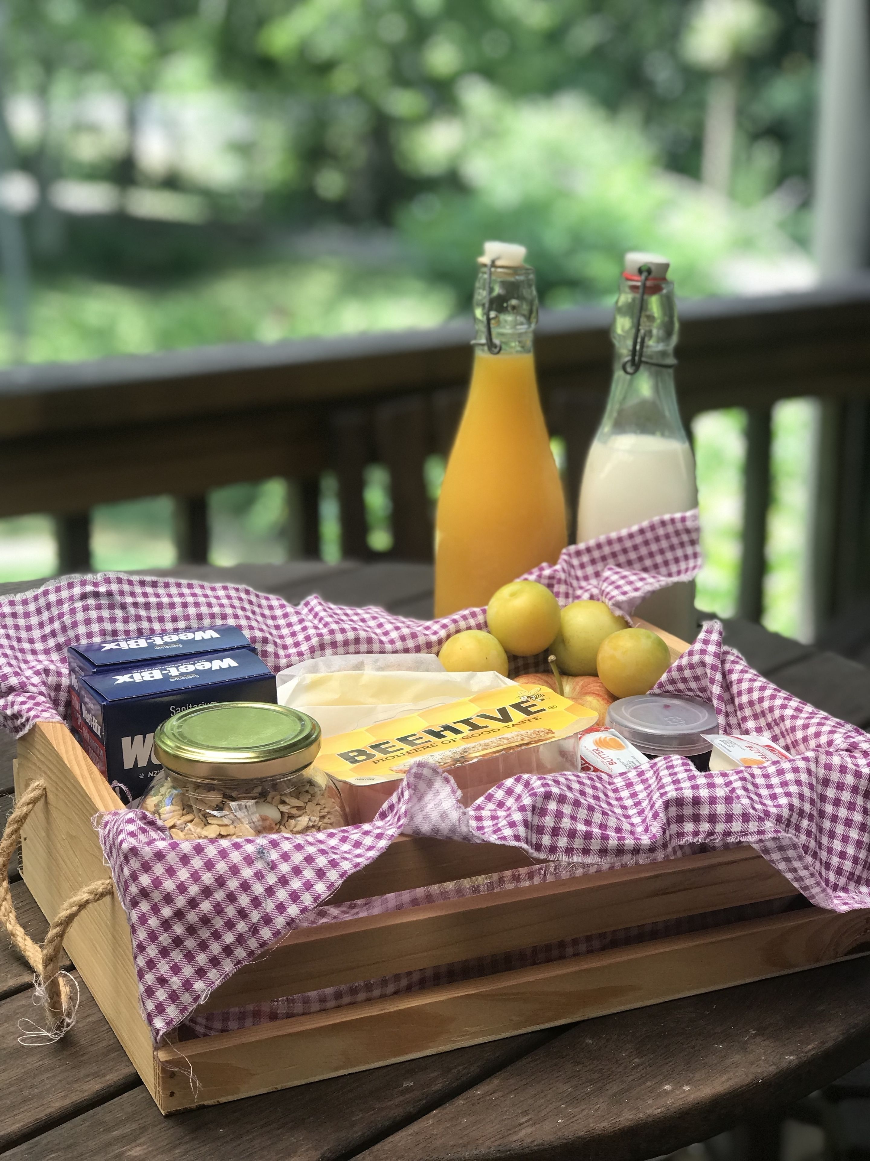 A wooden crate with a checkered cloth, containing jars, a loaf of bread, a bottle of orange juice, a bottle of milk, and a box of honey.