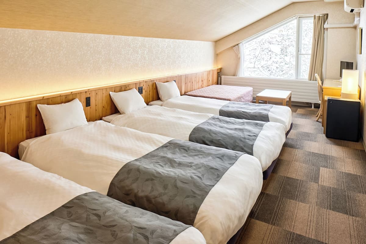 Une chambre d’hôtel avec quatre lits alignés contre un mur, une petite table et une fenêtre avec des rideaux.
