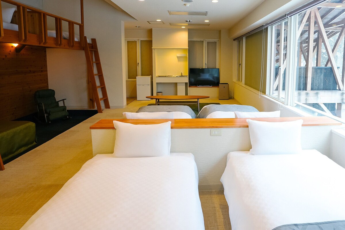 Habitación de hotel con dos camas, un televisor, una mesita y ventana con vista a árboles.