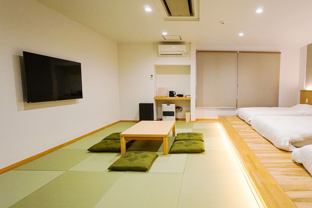 Habitación de hotel con televisión de pantalla plana, una mesa de madera, dos camas con ropa blanca y un sofá cama.