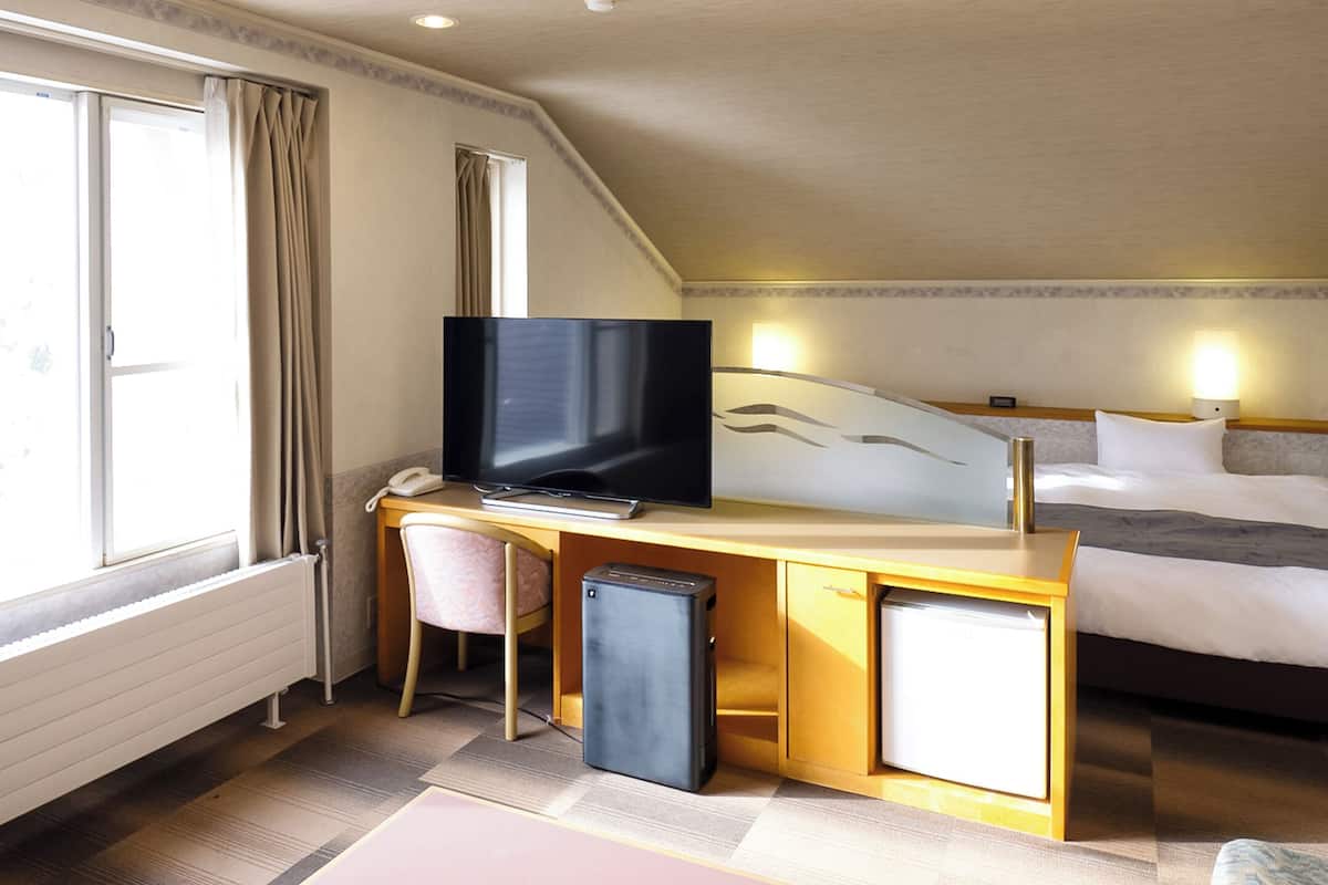 Une chambre d’hôtel comprenant un lit, un bureau avec un ordinateur et une télévision, un petit réfrigérateur et une fenêtre avec des rideaux.