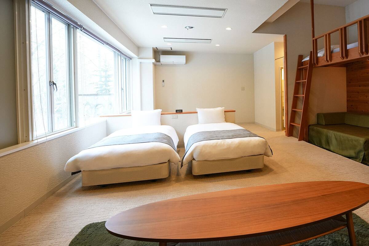 Une chambre avec deux lits, un lit superposé en bois, un canapé vert, une table en bois et de grandes fenêtres.