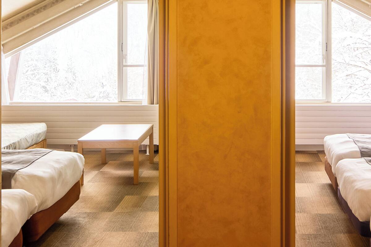 Une chambre avec deux lits, une petite table et de grandes fenêtres.