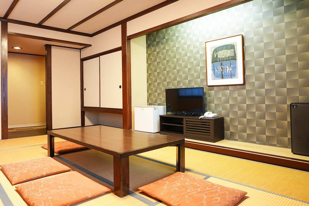 Une pièce de style japonais traditionnel, avec un sol recouvert de tatamis, une petite table en bois, des coussins, un téléviseur et un réfrigérateur.