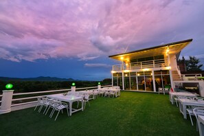 An outdoor patio with tables and chairs, a glass building with lights, and a view of the mountains. - Na Loei Boutique Resort Hotel (Mueang Loei)