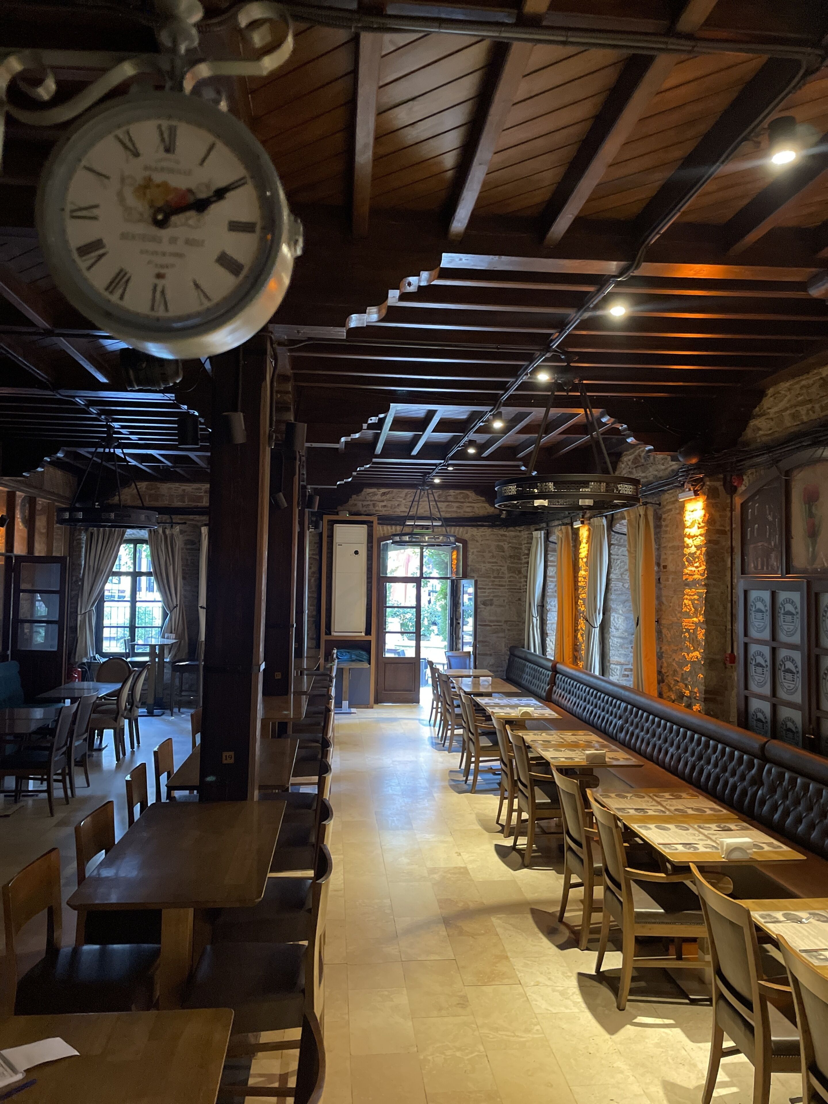 A restaurant with a long counter, wooden tables, and chairs. There is a clock hanging from the ceiling and a stone wall with framed pictures.