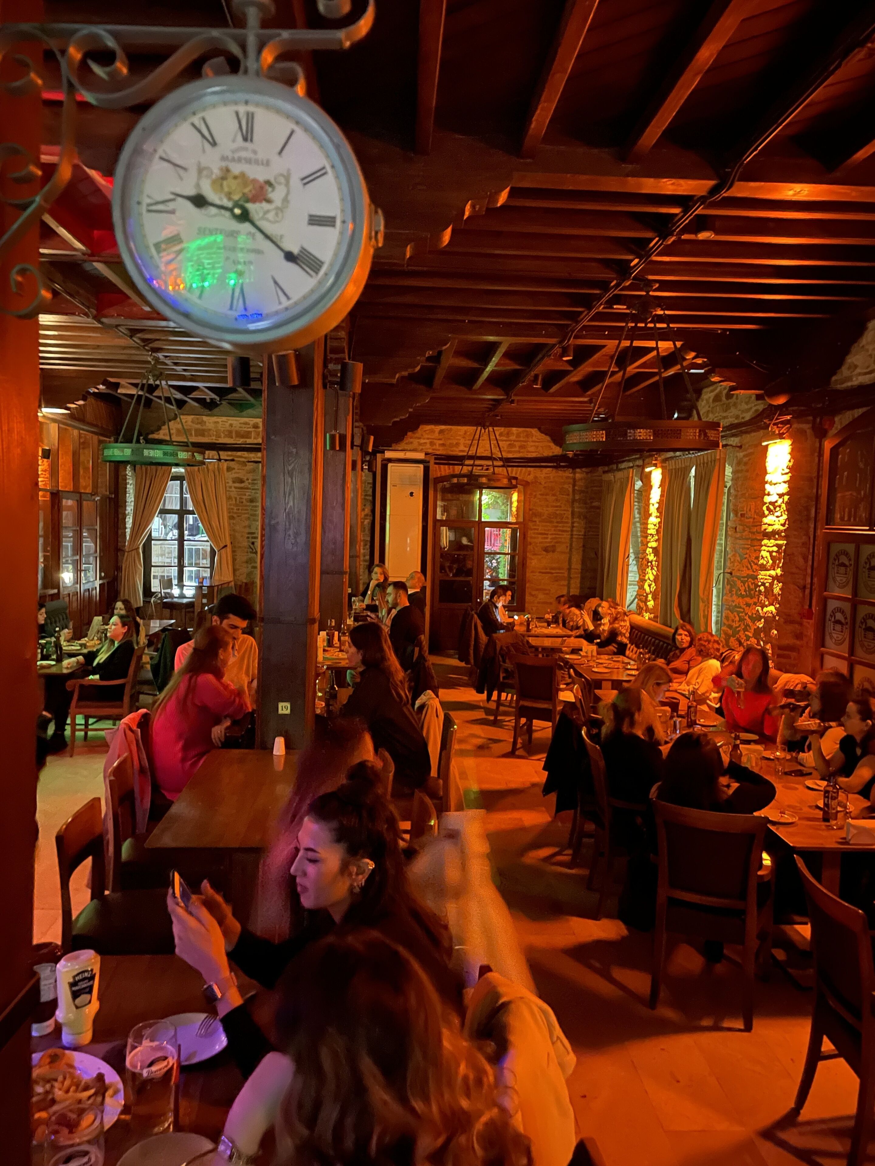 A restaurant interior with wooden beams, brick walls, and hanging lights. Tables are occupied by diners.
