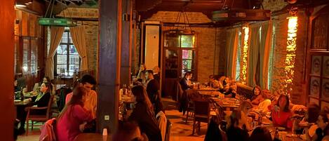 A restaurant interior with wooden beams, brick walls, and hanging lights. Tables are occupied by diners.