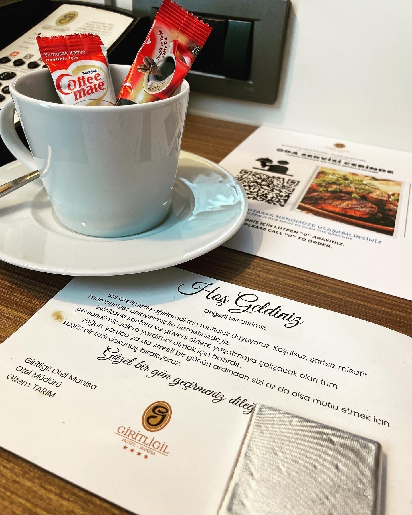 Quarto de hotel com uma chávena branca contendo café e um pacote de Nestle Coffee Mate. Há um menu e um código QR sobre a mesa.