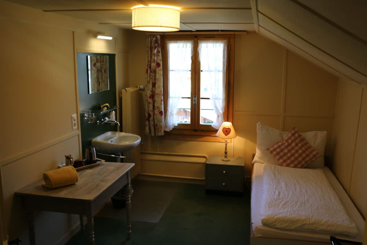 Un dormitorio pequeño con una cama individual, una mesita de noche, un pequeño aparador, una ventana con cortinas y un baño con lavabo y espejo.