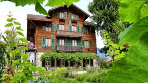 Un immeuble en bois à plusieurs étages, doté de volets verts, d’un balcon et d’un espace jardin aménagé avec des chaises.