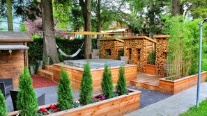 A wooden hot tub surrounded by raised planters with greenery and flowers, a hammock, and a wooden pergola.
