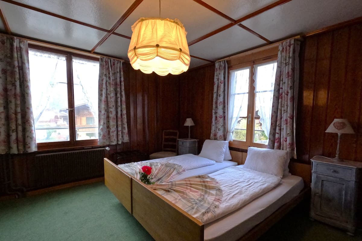 Un dormitorio con cama de madera, dos mesitas de noche, una mesita auxiliar, una lámpara y una ventana con cortinas.