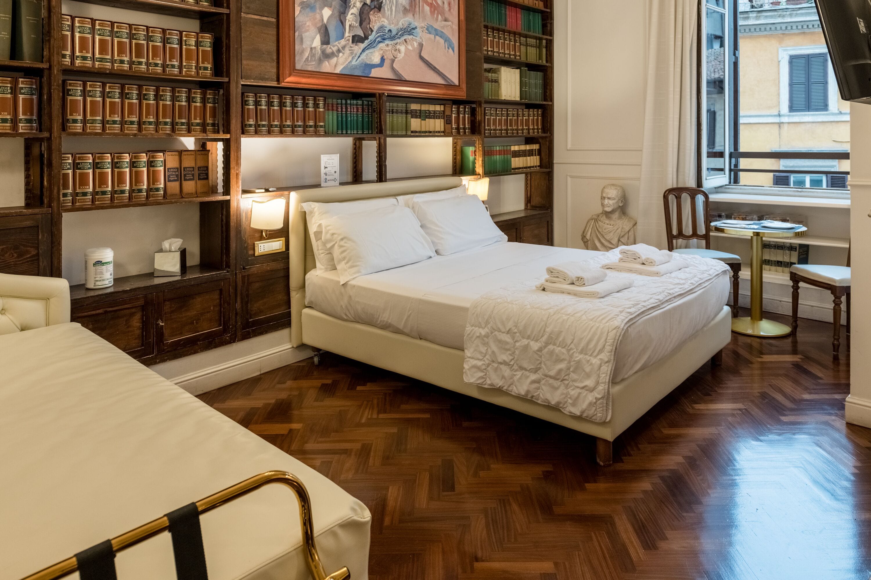 Una camera d'albergo con un letto grande, librerie in legno, uno scultura a busto e vista sulla città attraverso una finestra.