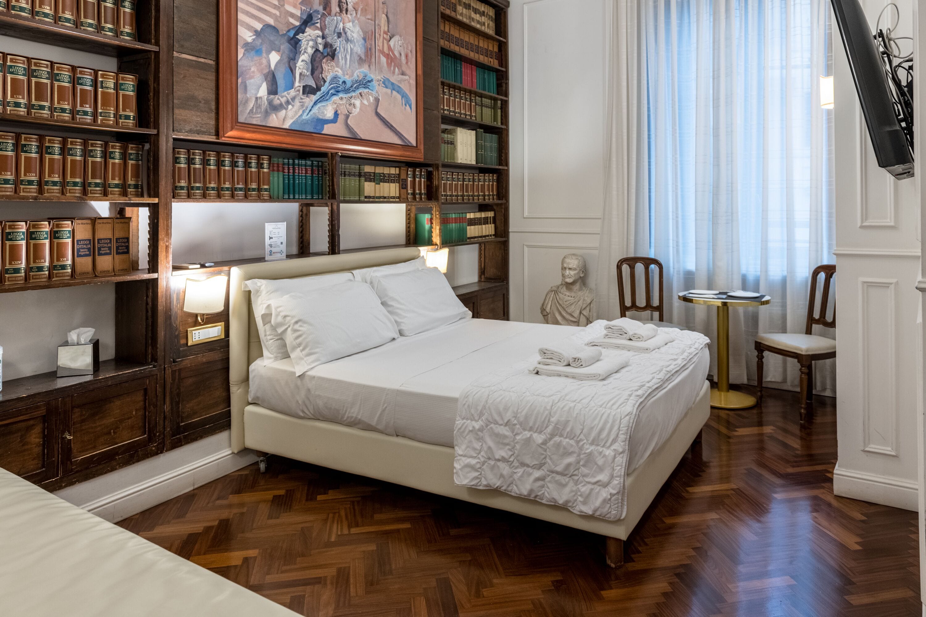 Camera d'albergo con un letto grande, pavimento in legno, una libreria piena di libri, un quadro e una statua.