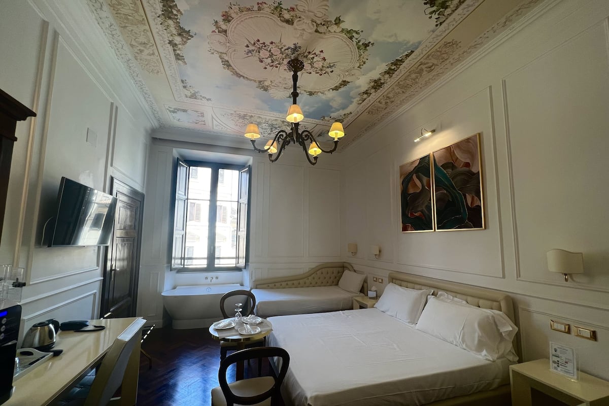 A hotel room with a bed, a desk, a chair, a chandelier, a window, and a painting on the wall.